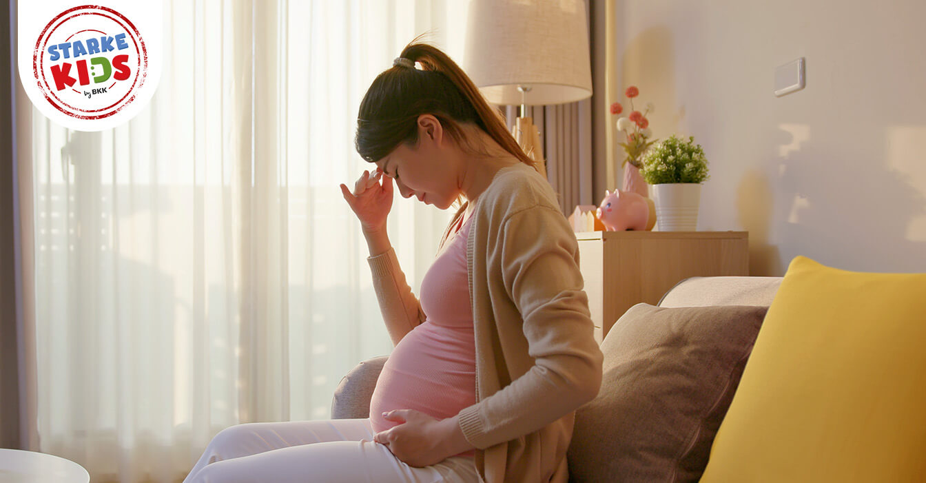 Schwangere Frau mit Kopfschmerzen sitzt auf dem Sofa, hält ihren Bauch und lehnt sich zurück, im sonnigen Zimmer mit weichen Farben.